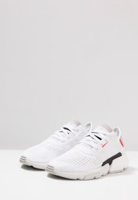 Zapatos deportivos blancos con parte superior de malla, acentos grises en el cuello y detalles negros en la suela. Presentan una textura perforada para mayor transpirabilidad.