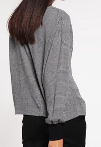 Donna che indossa una camicetta a maniche lunghe grigia e nera a scacchi con polsini neri, abbinata a pantaloni neri, vista di spalle.