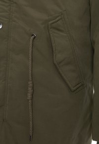 Olivegrüne Jacke aus strukturiertem Stoff, große Seitentasche und geflochtene Kordelzugdetails in der Taille mit metallenen Akzenten.