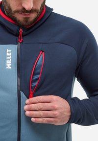 Granatowa i jasnoniebieska kurtka zapinana na zamek z czerwonymi akcentami. Posiada kieszeń zapinaną na zamek z siateczkową podszewką oraz dopasowany kaptur. Gładki, elastyczny materiał.