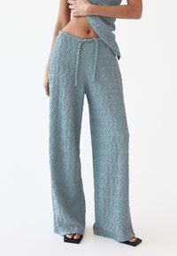 Light blue, textured wide-leg pants with a wavy pattern, featuring an elastic waistband and drawstring. Paired with black block-heeled sandals.