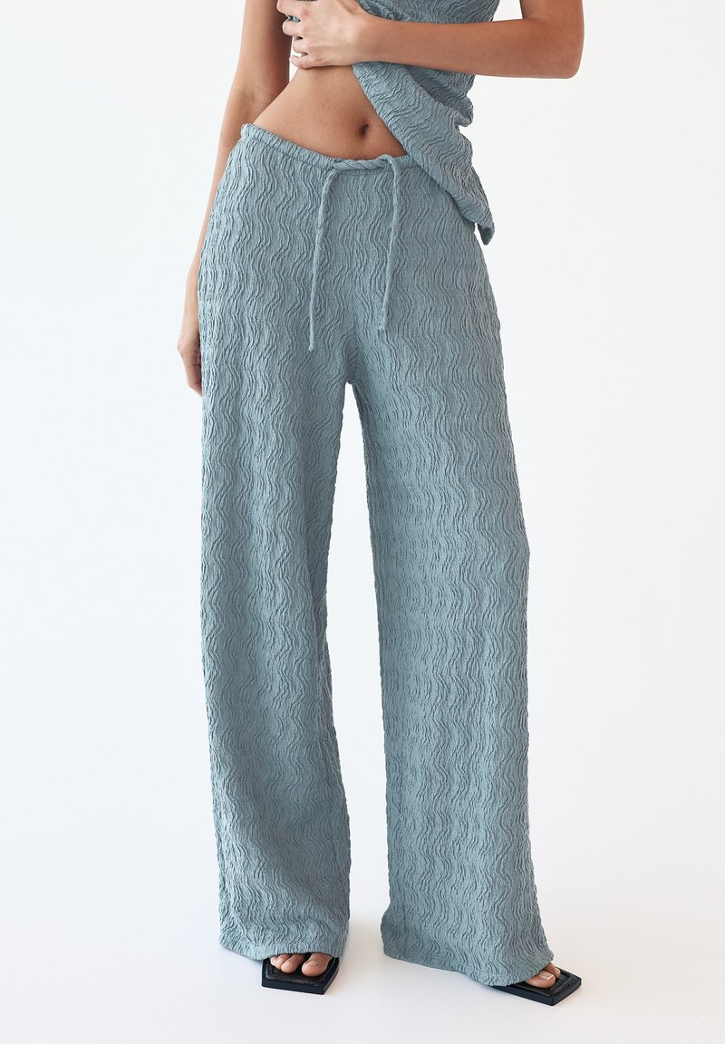 Light blue, textured wide-leg pants with a wavy pattern, featuring an elastic waistband and drawstring. Paired with black block-heeled sandals.