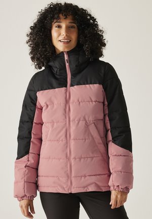 Donna con capelli ricci che indossa una giacca imbottita bicolore rosa e nera con cerniera, in piedi davanti a uno sfondo semplice.