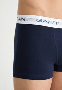 Marineblaue Boxershorts aus glattem Baumwollmaterial mit einem weißen Bund, auf dem das "GANT"-Logo in blauer Schrift steht. Nahtlose Seiten und Rücken.