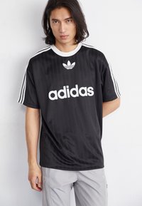 Fekete adidas póló fehér logóval és három csíkkal az ujjakon. Simább, könnyű anyagból készült, kerek nyakkivágással és finom függőleges mintázattal rendelkezik.