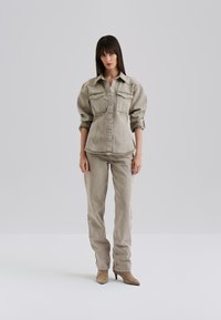 Beige denim takki, jossa on kaksi etutaskua ja napitus, yhdistettynä yhteensopiviin housuihin. Sujuva pinta ja vartalonmyötäinen muotoilu, esitettynä korkokenkien kanssa.