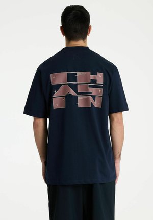 Man draagt een zwart T-shirt met grote abstracte blokletters in het bruin, gedrukt op de achterkant tegen een witte achtergrond.