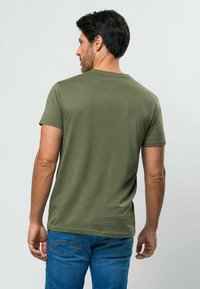 Alyvuogių žalios spalvos trumpomis rankovėmis medvilninė marškinėliai su klasikine apvaliomis iškirpte ir prie kūno prigludusia siluetu, derinami su mėlynais džinsais.