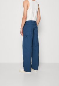 Henkilö, joka on pukeutunut löysiin sinisiin cargo-housuihin, valkoiseen tank-toppiin ja valkoisiin lenkkareihin, seisoo vaaleaa yksinkertaista taustaa vasten.