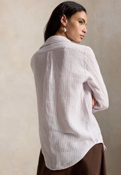 Femeie cu păr lung și închis la culoare, purtând o bluză roz deschis cu mâneci lungi cu dungi verticale subțiri și pantaloni maro, văzută din profil cu brațele încrucișate.