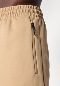 Calças de moletom bege feitas de um tecido macio, com um cós elástico e um bolso lateral com zíper. Textura suave com detalhes mínimos.