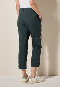 Donkergroene cropped cargobroek met een zijritszak, knoopzakken op de achterkant en een gladde textuur, gecombineerd met witte sneakers.