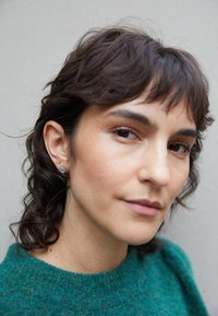 Suéter verde texturizado, cabelo castanho ondulado com franjas curtas, apresentando um pequeno brinco de metal. Características faciais suaves e neutras contra um fundo claro.