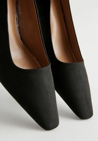 Zapatos de tacón alto de ante negro con punta afilada y un interior de cuero suave, que presentan un diseño minimalista y una silueta elegante.