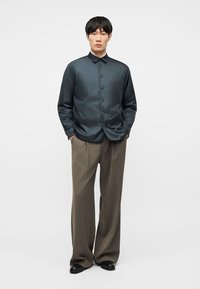 Jeune homme debout, les mains dans les poches, portant une chemise sombre à boutons et un pantalon large marron, sur un fond blanc uni.