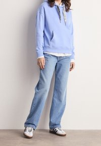 Person som bär en ljusblå hoodie, vit skjorta under, lösa blå jeans och vita sneakers med svarta ränder står inomhus.