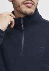 Suéter de forro polar azul marino con cuello alto, con cierre frontal de cremallera y un pequeño logo en el pecho. Textura suave, diseño ligero.