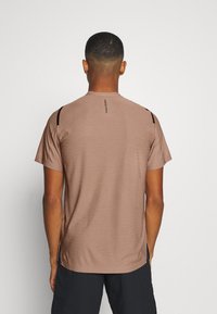 Camiseta atlética de manga corta color marrón. Presenta un material texturizado con líneas sutiles, acentos negros en los hombros y un logotipo en la parte posterior.