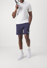 Polo blanc, short bleu marine avec des bandes latérales, baskets blanches, tenant une raquette de tennis. Le design est sportif avec une coupe décontractée.
