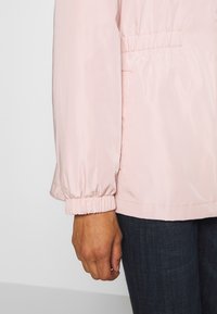 Ljusrosa, nylonjacka med elastiska ärmslut, innyttjad midja och sidofickor. Tyget har en slät, lätt glänsande yta.