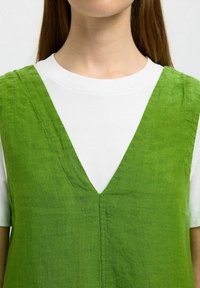 Vestido de lino verde con un profundo escote en forma de V, superpuesto a una camiseta blanca. La tela tiene una apariencia texturada y arrugas naturales.