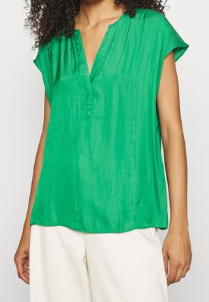 Vrouw draagt een mouwloze felgroene blouse met een V-hals, gecombineerd met een witte broek met hoge taille, staand tegen een effen achtergrond.