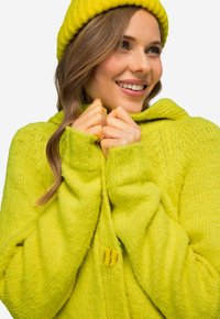 Světle žlutý pletený svetřík s kapucí, vykazující hrubou strukturu, žebrované manžety a velké žluté zapínání na knoflík.