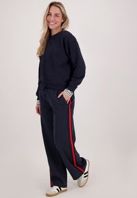 Marineblauwe sweatshirt met geribbelde mouwen over gestreepte marineblauwe broek met rode zijkanten, gedragen met witte sneakers met zwarte accenten.