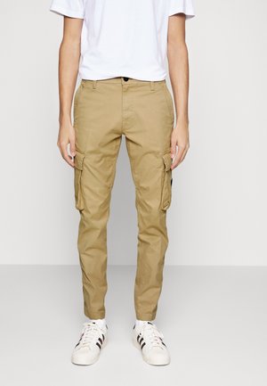 Osoba w beżowych spodniach typu cargo z bocznymi kieszeniami, białych trampkach z czarnymi paskami oraz gładkiej białej koszulce na tle o neutralnym kolorze.