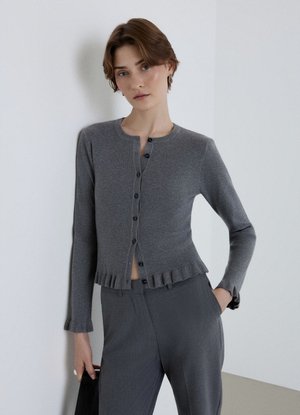 Giovane donna con i capelli corti che indossa un cardigan grigio a coste aderente e pantaloni grigi abbinati, in piedi contro un muro chiaro e semplice.