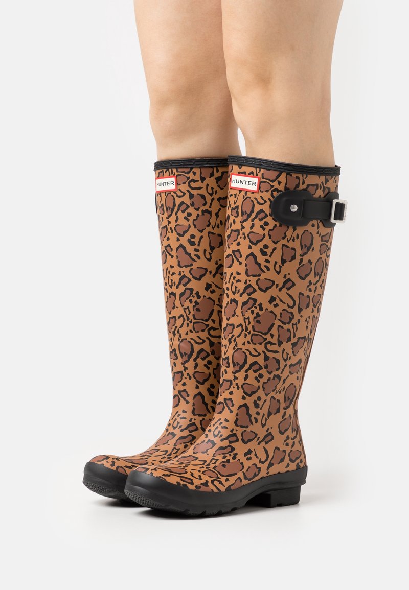Jambes portant des bottes de pluie marron montantes jusqu'aux genoux avec des taches léopard noires, des semelles noires et une sangle noire réglable avec boucle sur le côté.