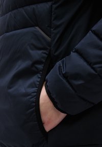 Granatowa kurtka puchowa z zamykaną kieszenią na zamek. Posiada miękką fakturę, pikowany wzór oraz dopasowanie zapewniające bezpieczeństwo wokół mankietów i dołu kurtki.