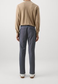 Beżowa koszula z długim rękawem w połączeniu z ciemnoszarymi spodniami i białymi butami, pokazująca prosty, swobodny krój od tyłu.