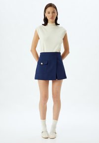 Navyblauwe mini-jurk met een zijflapzak, gouden knoopdetails en een gestructureerde pasvorm, gecombineerd met een crèmekleurige mouwloze top en lichte sokken.
