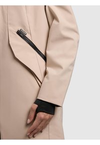 Bež rokav plašča s črnim manšetnim odprtinim za palec in poševnim črnim žepom z zadrgo ter majhno belo logotipsko etiketo blizu zapestja.