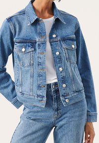 Ljusblå denimjacka med en klassisk design, två bröstfickor, silverknappar och kontrastsömmar. Bärs över en vit skjorta.