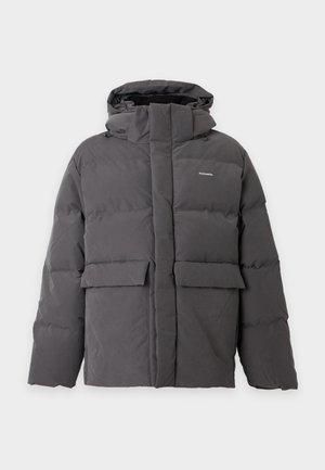 Un homme porte une doudoune à capuche gris foncé, une chemise bleue, un pantalon beige et des chaussures foncées sur un fond clair uni.