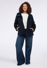 Giacca in misto lana blu navy con colletto alto, due tasche frontali e bottoni con dettagli dorati. Abbinata a una camicia di colore chiaro e jeans a gamba larga.