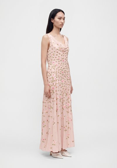 Femme debout portant une longue robe rose sans manches avec broderie florale et chaussures pointues rose clair sur un fond uni.