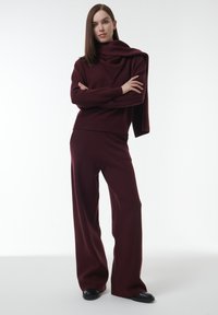 Bordeaux gebreide outfit met een hoge hals trui en wijde broek, voorzien van een gedrapeerde sjaal en zwarte schoenen, tegen een witte achtergrond.