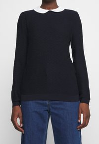 Marinblå långärmad tröja i texturerad tyg, vit Peter Pan-krage och ribbade muddar och nederkant. Matchad med blå denimbyxor.