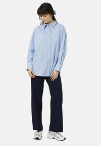 Chemise boutonnée bleu clair et blanche à rayures avec un col, portée avec un pantalon marine et des baskets argentées. Le tissu semble lisse, avec une coupe décontractée.