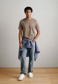 Καφέ εφαρμοστή μπλούζα, ανοιχτόχρωμο τζιν, καρό πουκάμισο δεμένο στη μέση και λευκά αθλητικά παπούτσια. Το φόντο είναι απλό και ανοιχτόχρωμο.