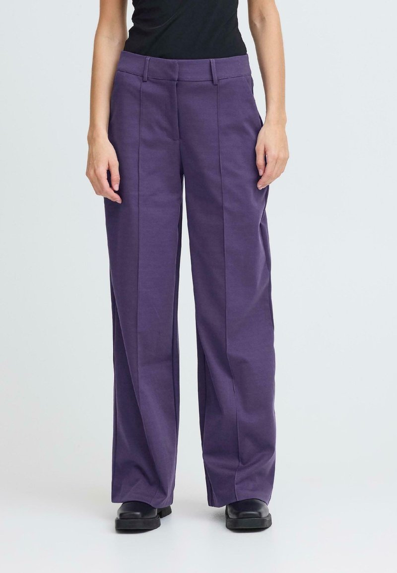 Pantalons évasés en coton violet, avec des plis sur le devant et une taille plate. Associés à des chaussures noires, sur un fond uni.