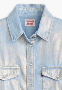 Camicia in denim azzurro chiaro con finitura metallica, chiusura a scatto, due tasche frontali e colletto. Presenta l'etichetta Levi's sul colletto.