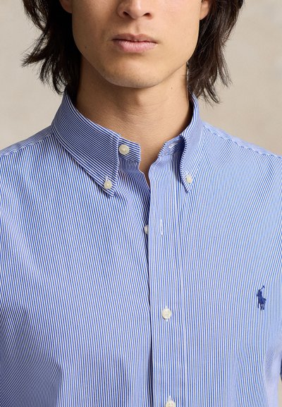Férfi kék-fehér csíkos, gombos galléros ingben, a gallér begombolva, és a mellkason hímzett logóval.