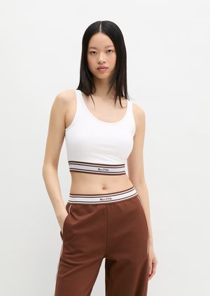 Tânără cu păr negru drept poartă un top alb scurt și pantaloni maro, stând cu o mână în buzunar, pe un fundal simplu.