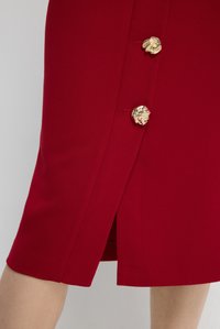 Jupe rouge en tissu texturé, dotée d'une fente sur le côté et de trois boutons décoratifs en or avec des motifs intriqués à l'avant.