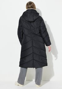 Giacca a vento nera con cappuccio, caratterizzata da un design a zigzag e un colletto interno morbido. Il modello indossa pantaloni larghi grigi e stivali color crema.
