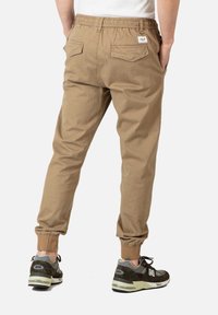 Tan joggings met een elastische tailleband, taps toelopende pijpen en twee achterzakken. Draagt een paar donkere sneakers met een grijs en zwart kleurenschema.